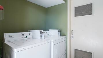 Un área de lavandería con dos lavadoras y secadoras apiladas, una pared verde y una puerta con rejilla de ventilación.
