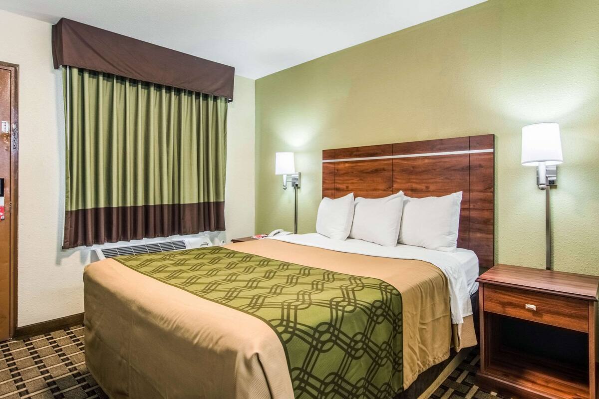 Quarto de hotel com cama, mesa de cabeceira, luminária, janela com cortinas e ar-condicionado fixado na parede.
