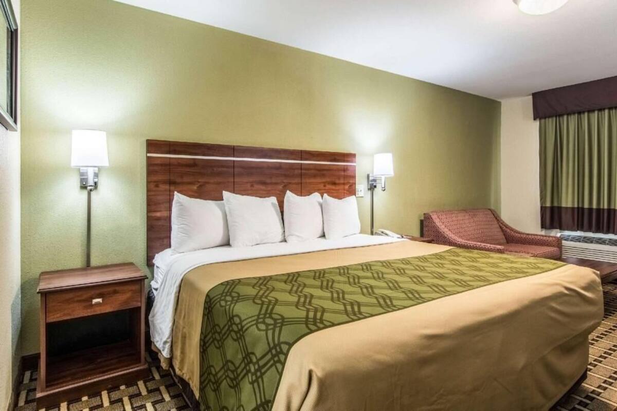 Quarto de hotel com cama, duas luminárias de cabeceira, uma cadeira, mesa de cabeceira e uma janela com cortinas.