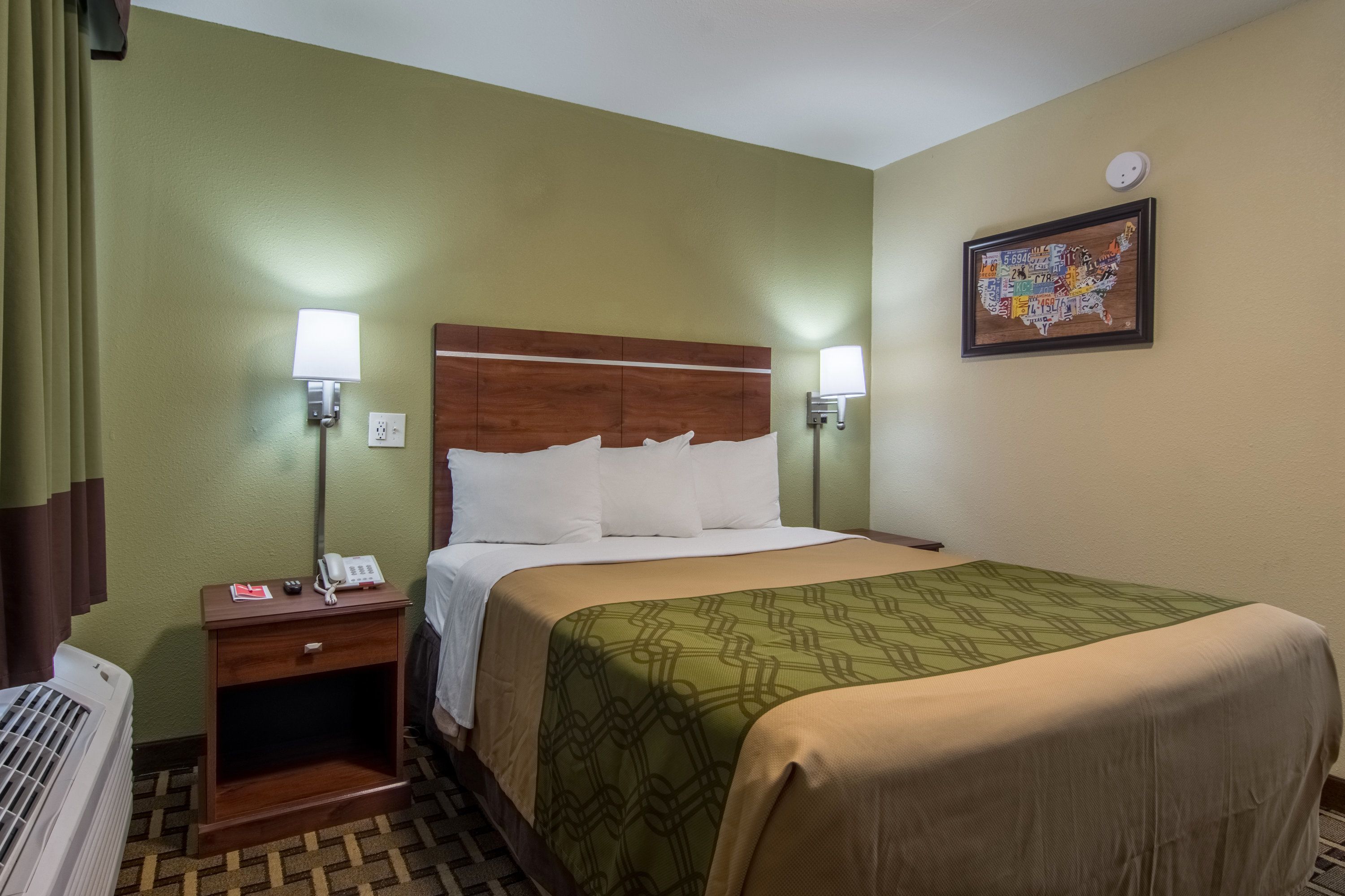 A hotel room with a bed, bedside lamps, a map on the wall, and a small bedside table with a phone and a book.