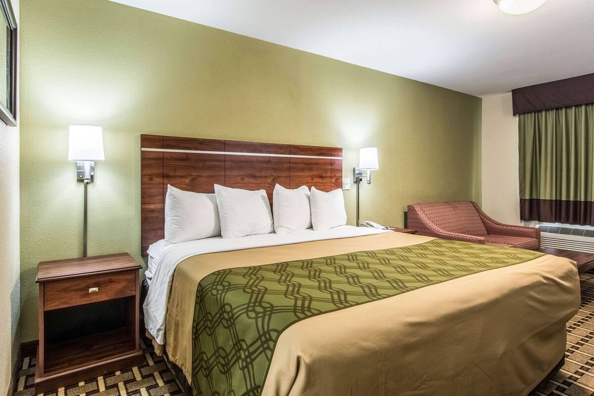 Quarto de hotel com cama, mesa de cabeceira, cadeira e janela com cortinas.