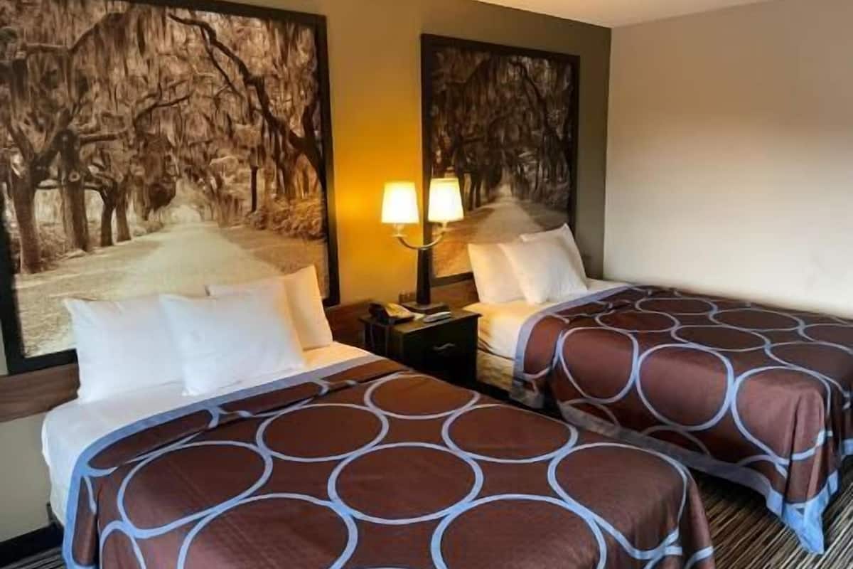 A hotel room with two beds, a nightstand, a lamp, and wall art depicting trees.