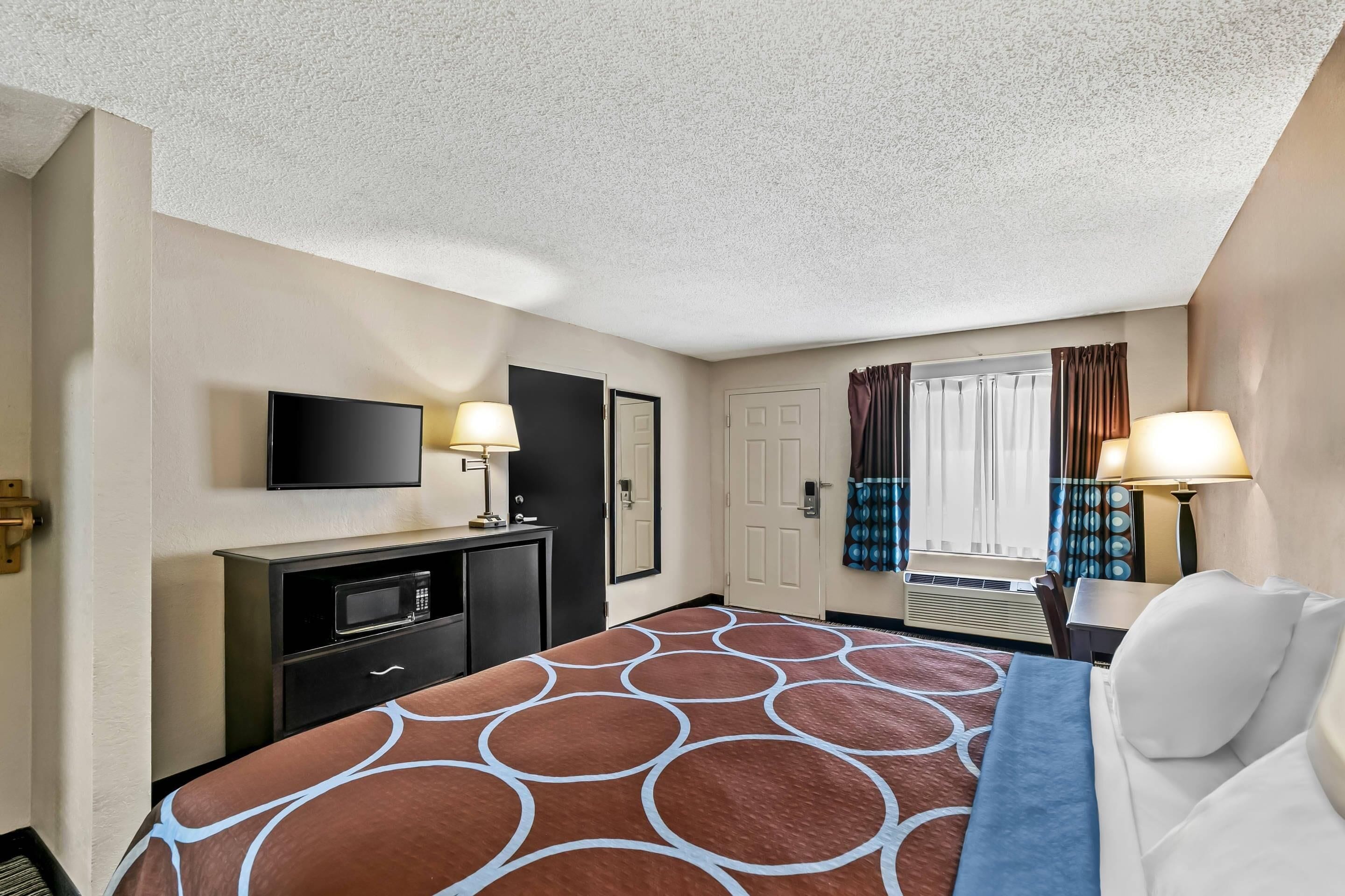 A hotel room with a bed, a flat-screen TV, a desk with a lamp, a microwave, and a window with curtains.