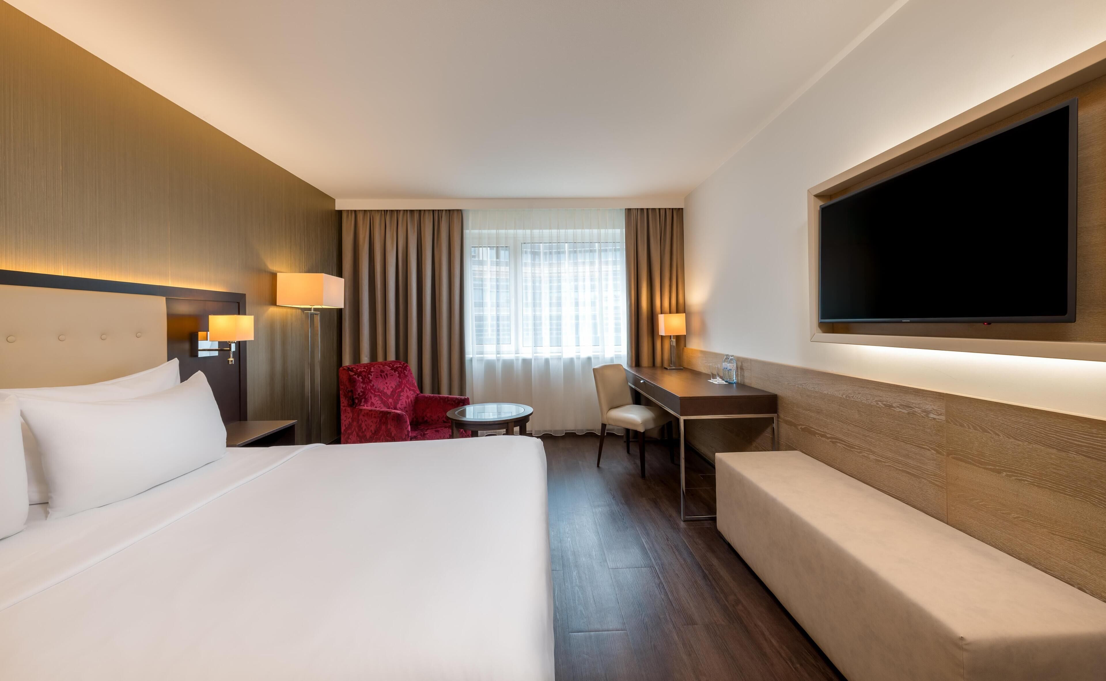 Habitació d'hotel amb un llit gran, un escriptori, una cadira vermella, un sofà i una televisió penjada a la paret.
