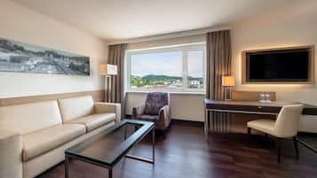 A hotel room with a sofa, armchair, desk, and a large window with a view of a cityscape.