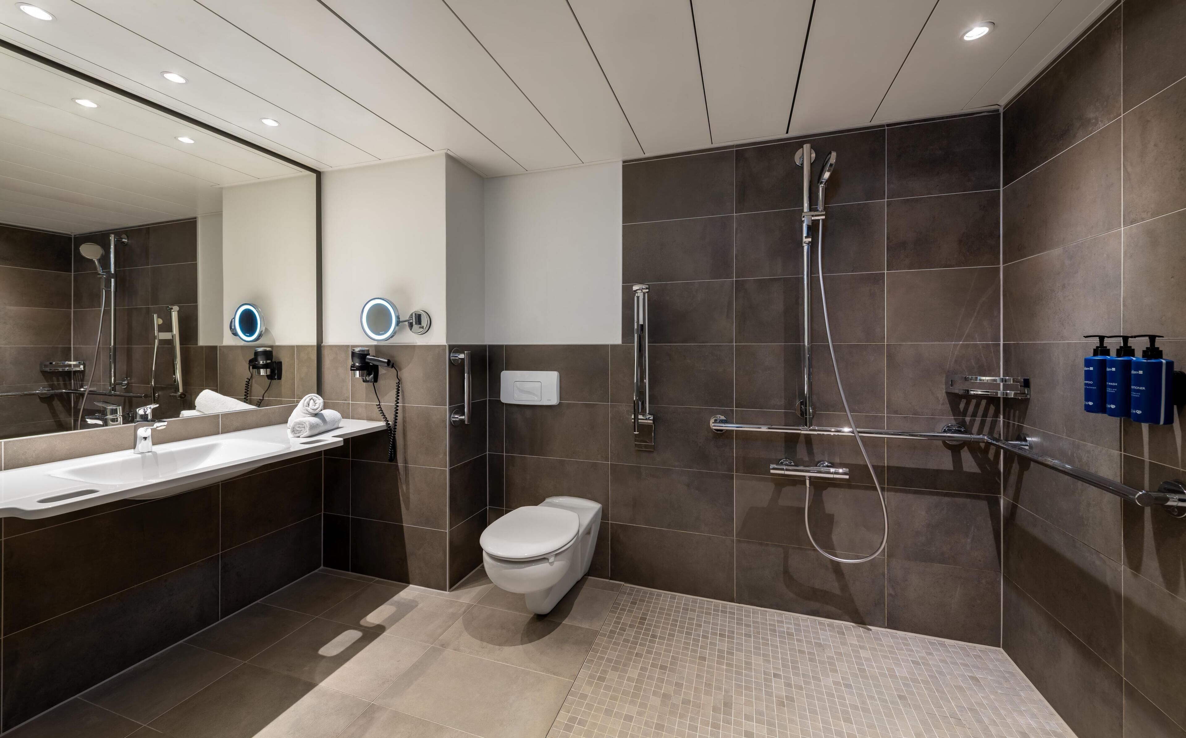 Un baño moderno con un espejo grande, ducha, inodoro y tocador con lavamanos.