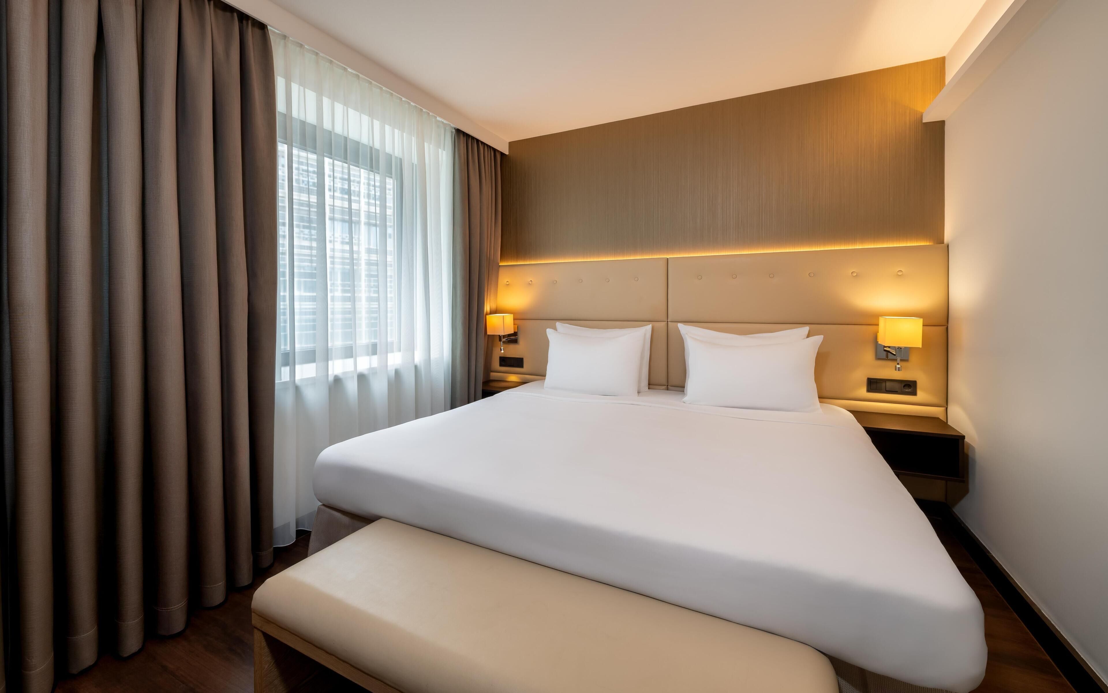 A hotel room with a large bed, a bench, two bedside lamps, and a view of a building through sheer curtains.