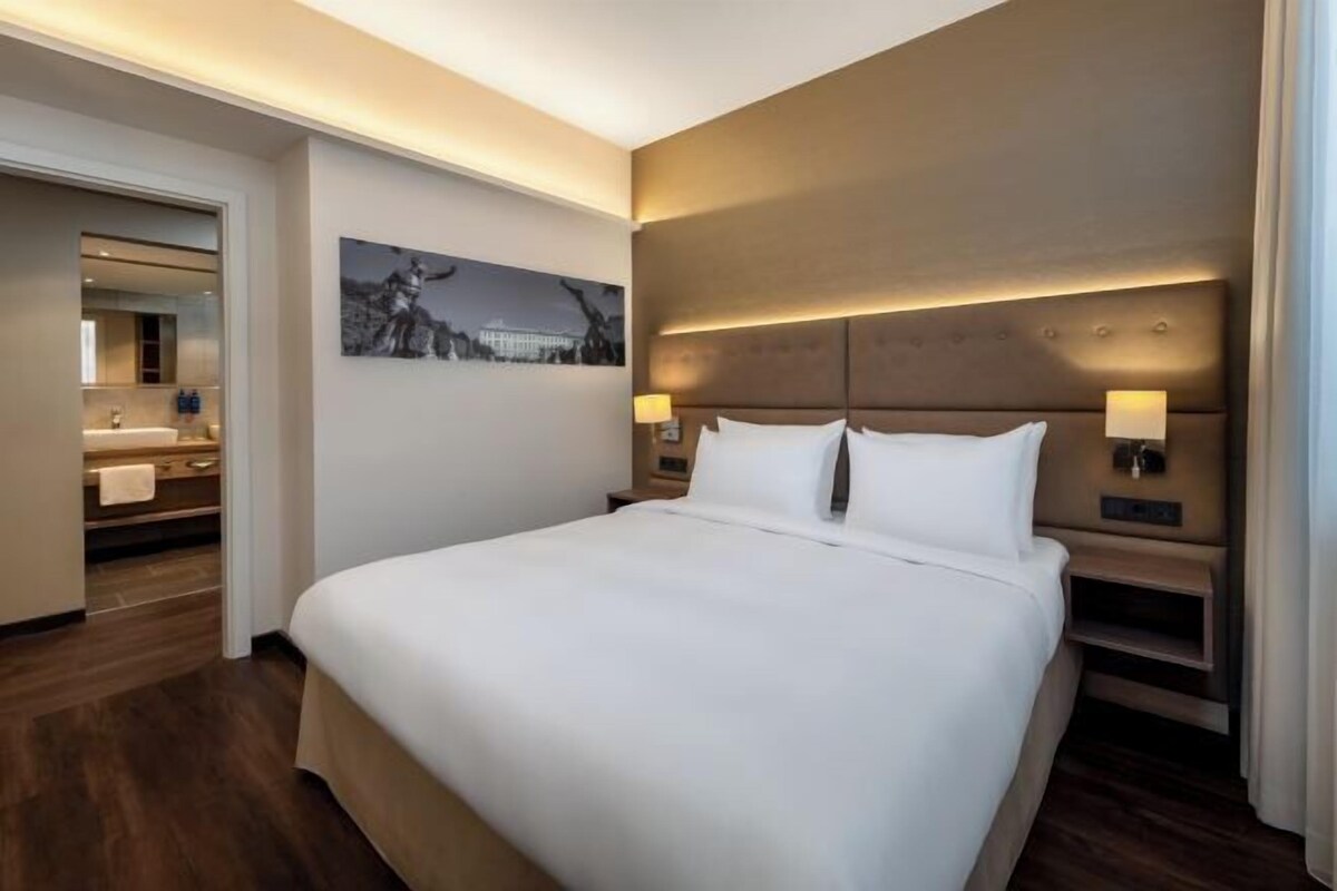 Ein Hotelzimmer mit einem großen Bett, Nachttischlampen, einem Kopfteil mit integrierter Beleuchtung, einem gerahmten Bild an der Wand und einem durchgegangenen Tür einsehbaren Badezimmer.