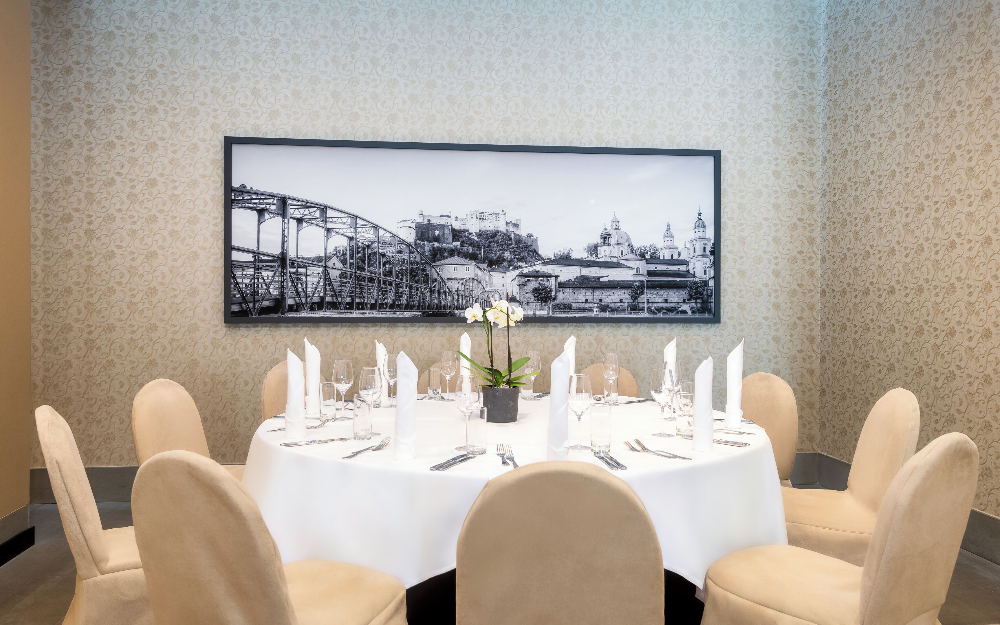 Une table à manger dressée pour six personnes, avec des nappes blanches, des chaises et un tableau représentant un paysage urbain accroché au-dessus.