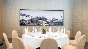 Une table à manger dressée pour six personnes, avec des nappes blanches, des chaises et un tableau représentant un paysage urbain accroché au-dessus.