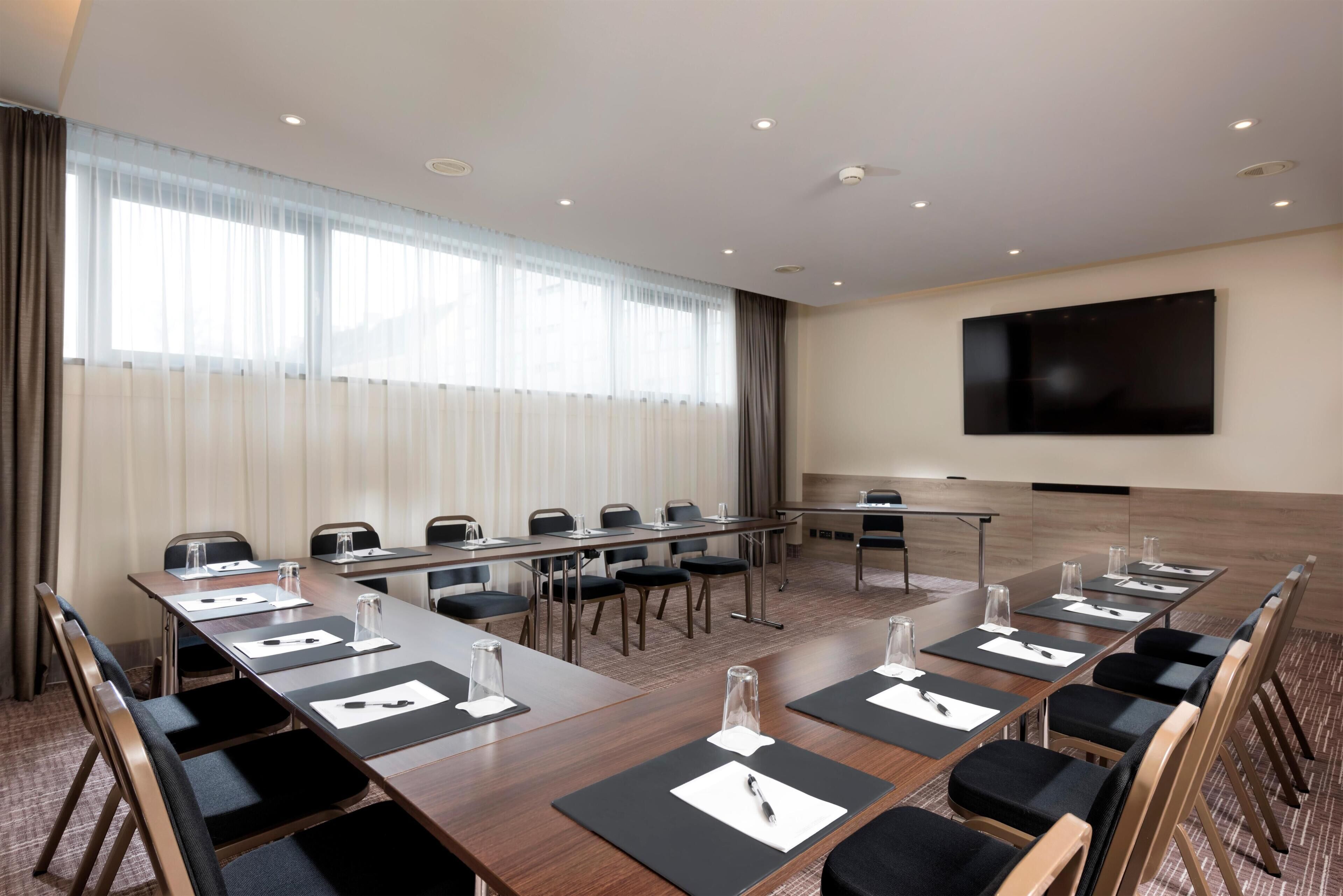 A conference room with a long table, chairs, a flat-screen TV, and a large window with curtains.