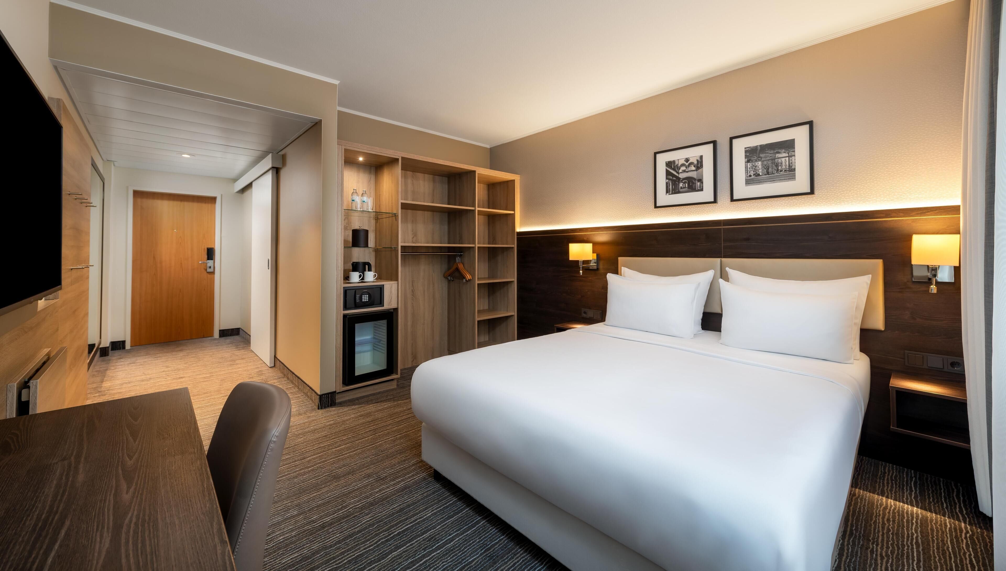 A hotel room with a bed, bedside tables, a desk, a chair, a wardrobe, and a television.