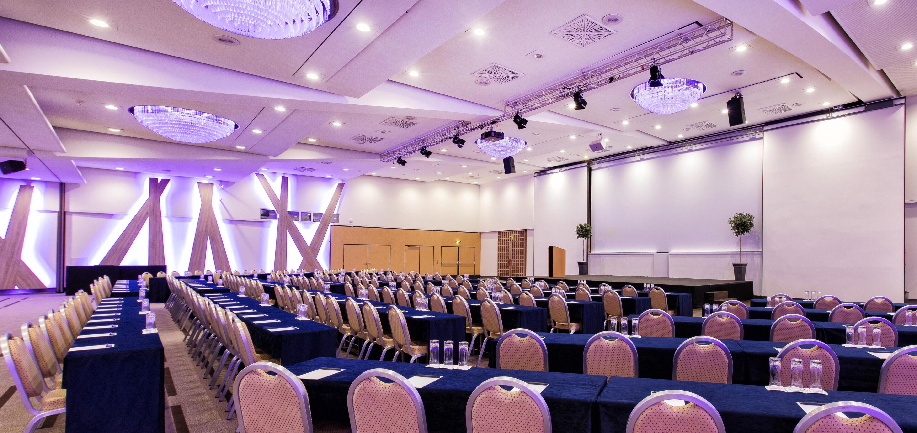 Un salón de conferencias con mesas y sillas dispuestas en filas, un escenario y una pantalla grande.