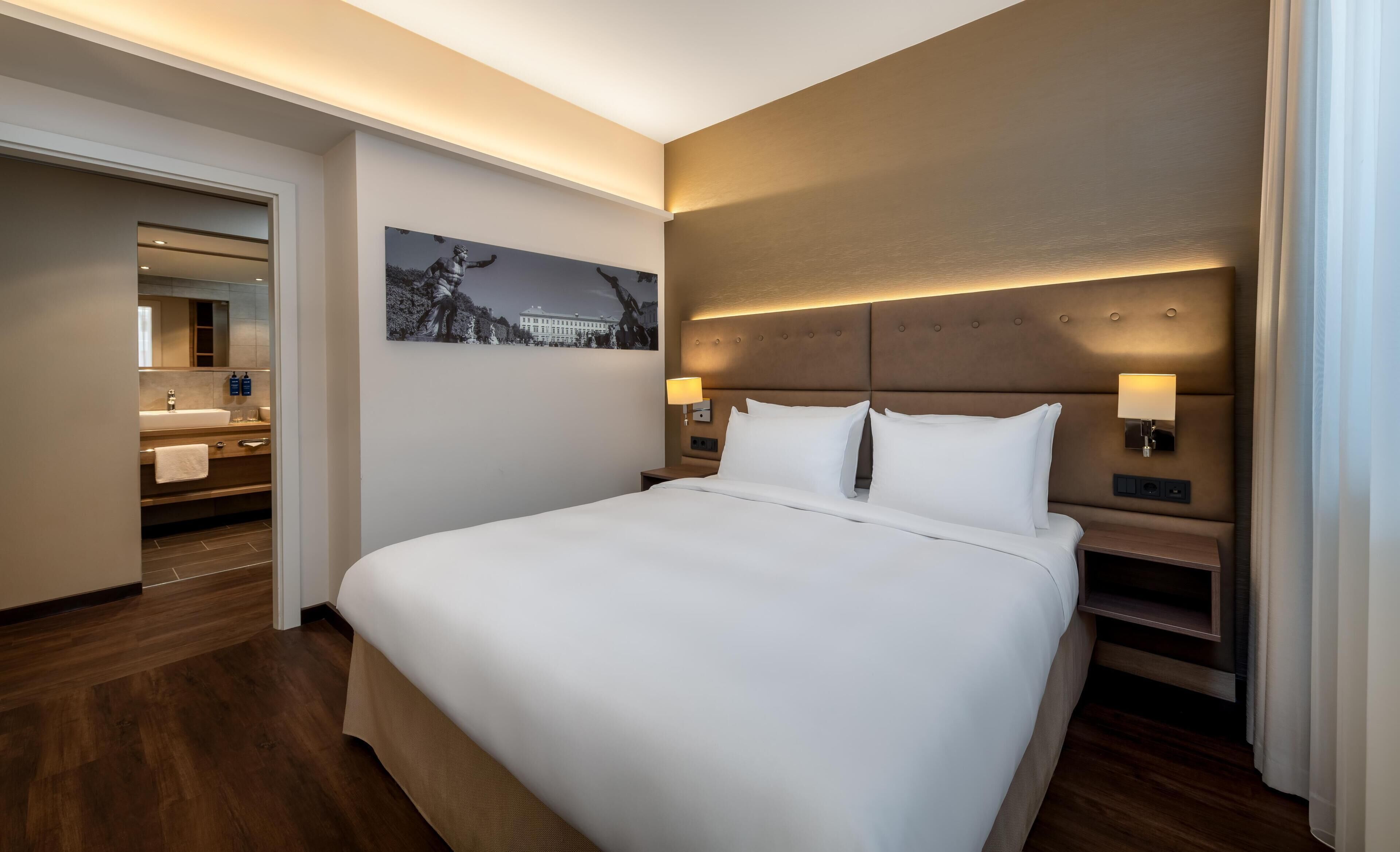 A hotel room with a large bed, a nightstand, a wall-mounted lamp, and a painting.