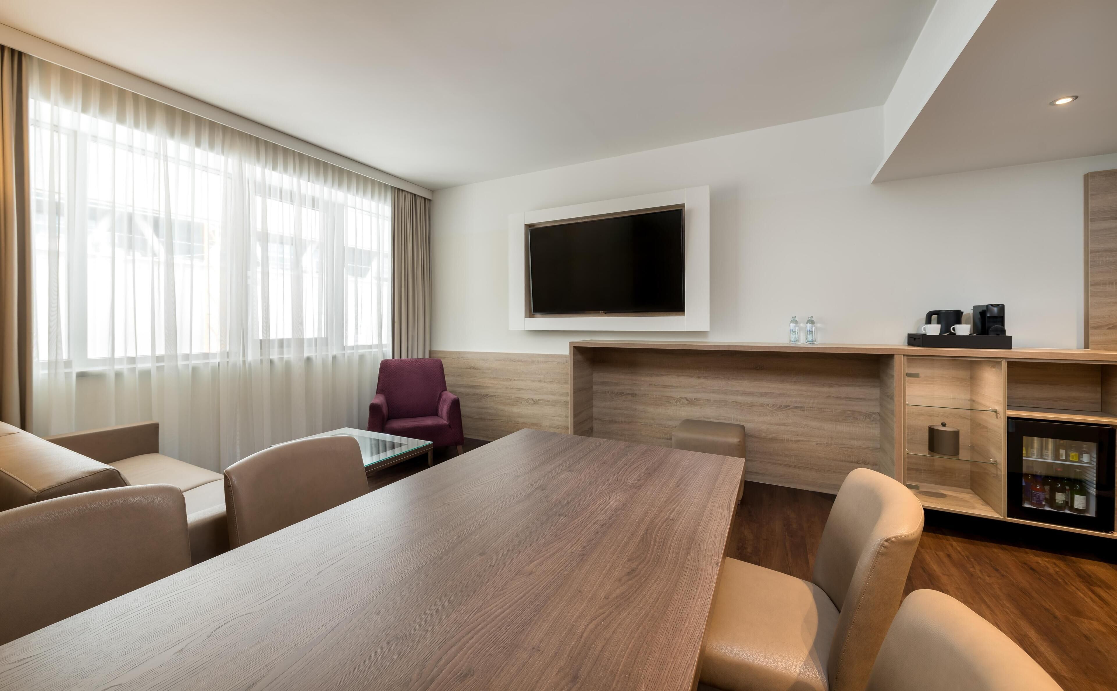 Quarto de hotel com uma grande mesa de madeira, televisão de tela plana na parede, área de estar com cadeiras e um minibar.