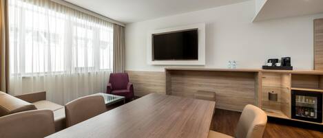 Una habitación de hotel con una amplia mesa de madera, un televisor de pantalla plana montado en la pared, una zona de estar con sillas y un minibar.