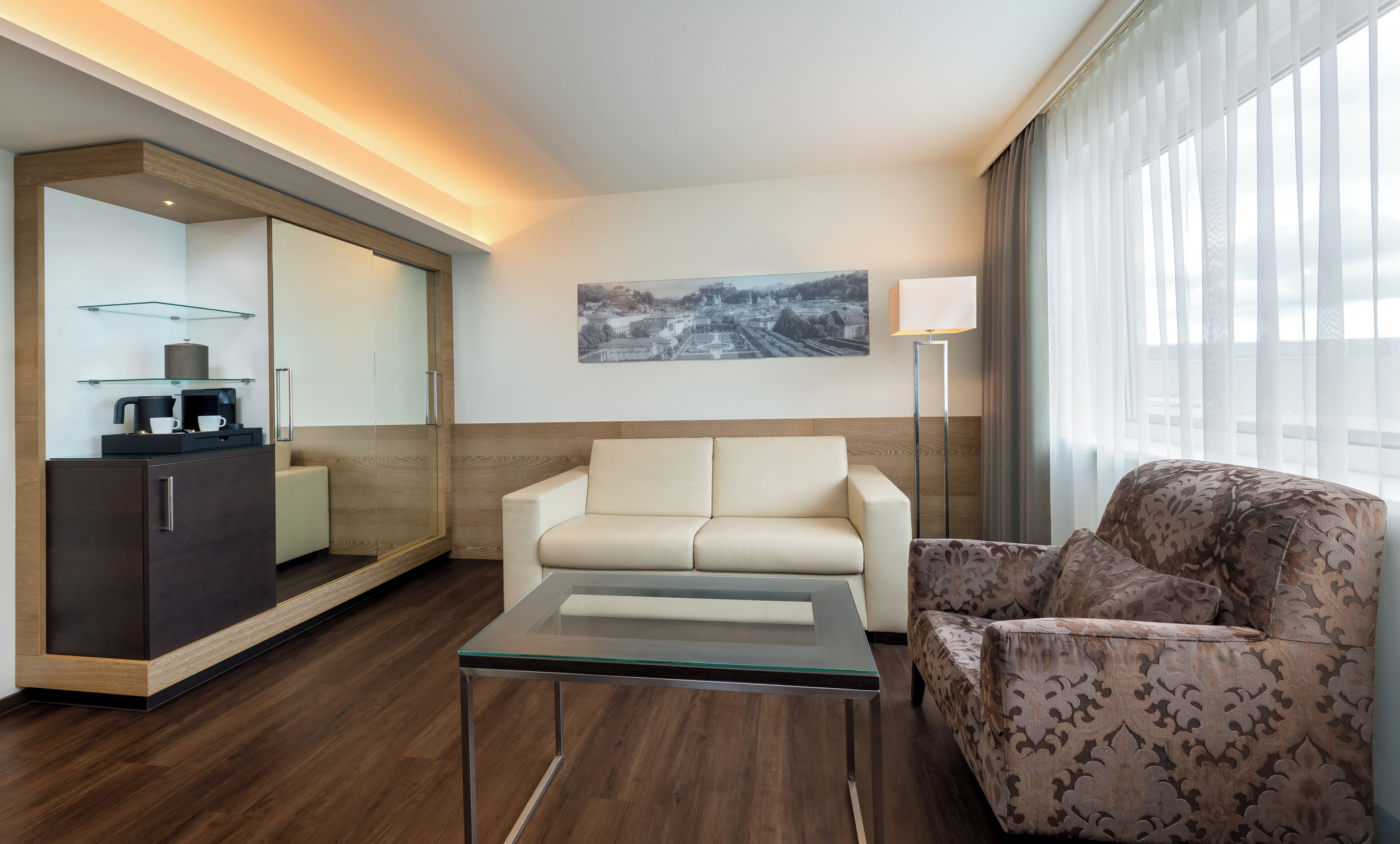 Quarto de hotel moderno com sofá, poltrona e mesa de centro de vidro.