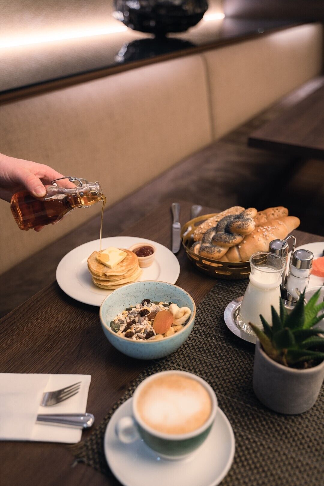 A breakfast setting with pancakes, a bowl of cereal, a cup of coffee, and a bottle of syrup being poured.