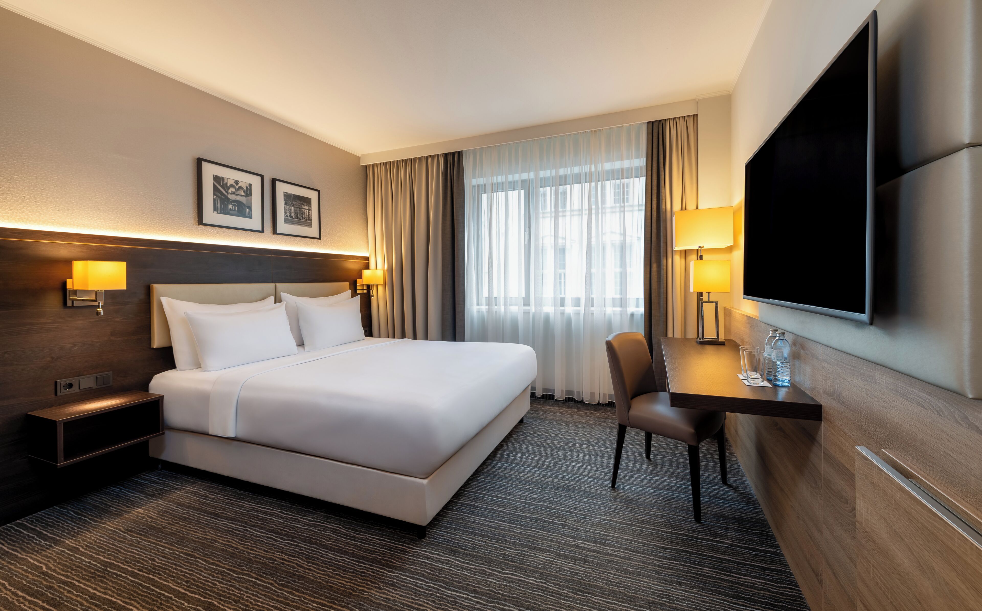 A hotel room with a bed, bedside lamps, a desk with a chair, a flat-screen TV, and a large window with curtains.