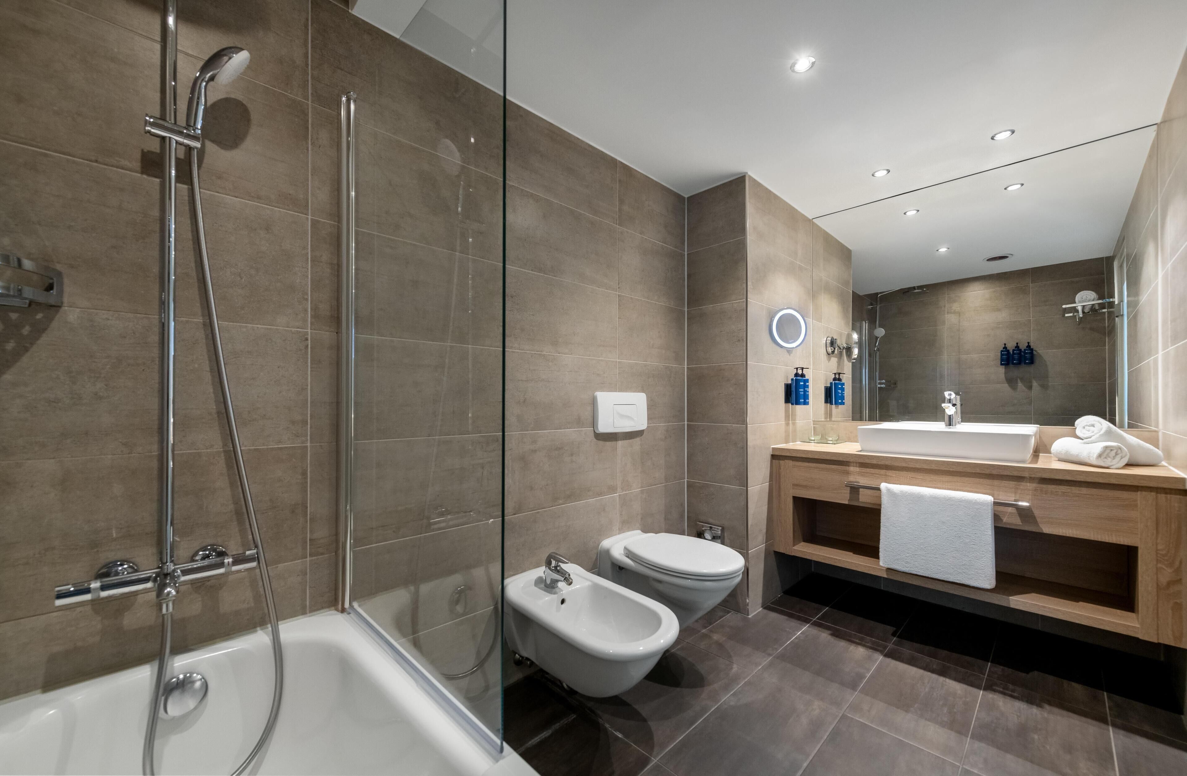 Un baño moderno con ducha envuelta en vidrio, una bañera independiente, un bidé y un tocador de madera con espejo y perchero.