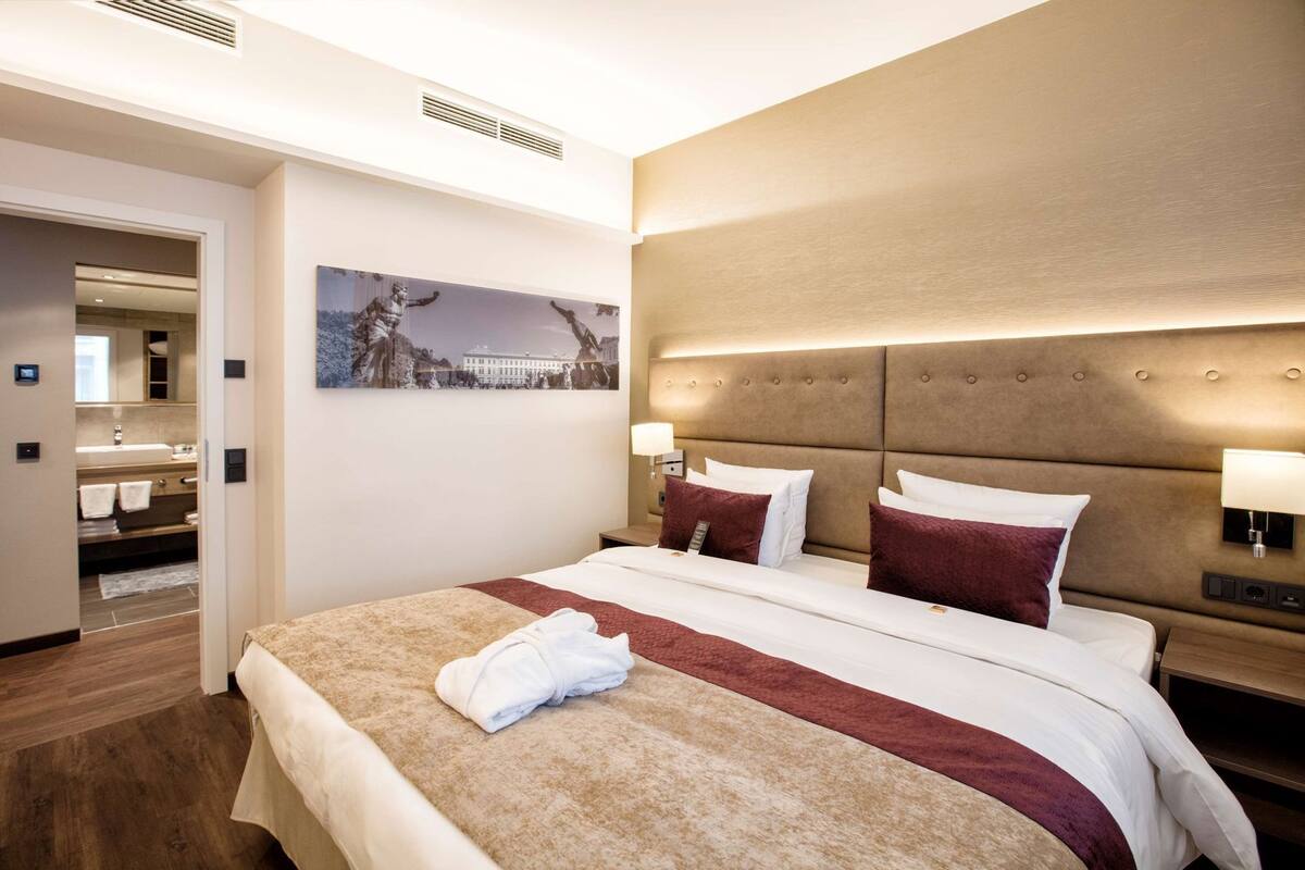 A hotel room with a large bed, a headboard with integrated lighting, a nightstand with a lamp, and a painting on the wall.