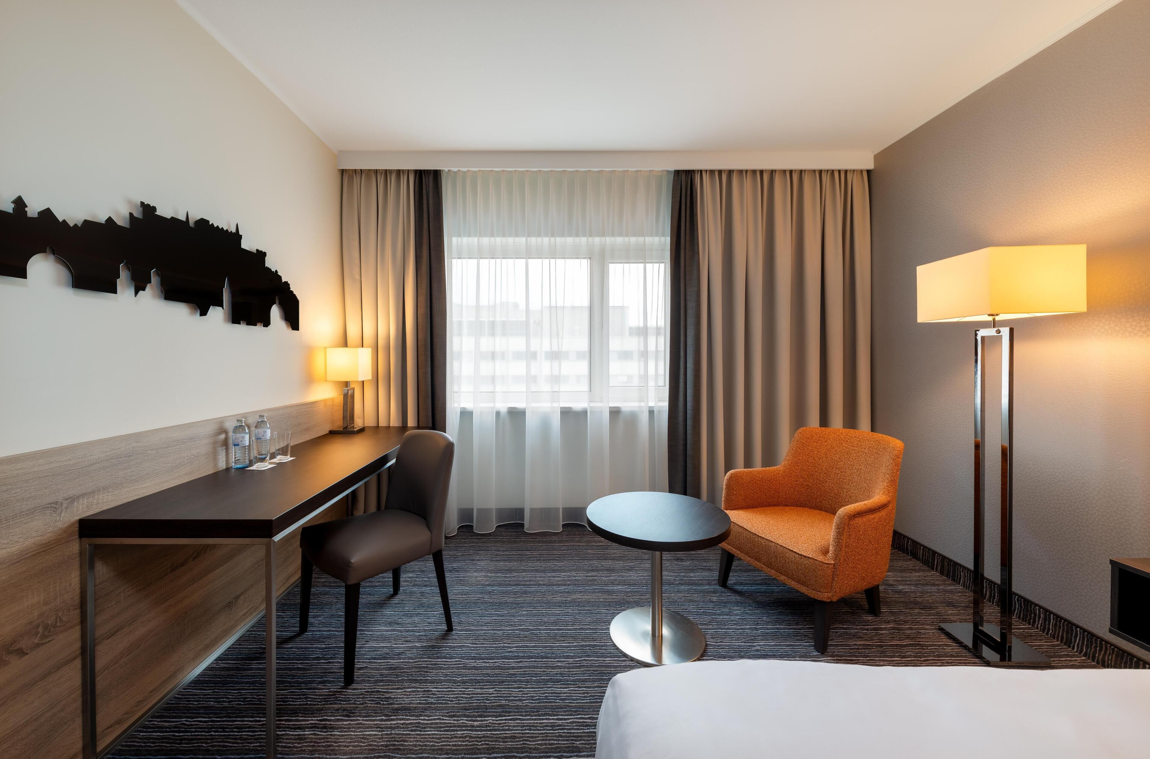 Habitación de hotel con cama, escritorio, silla, una mesita redonda, lámpara y luz empotrada en la pared.