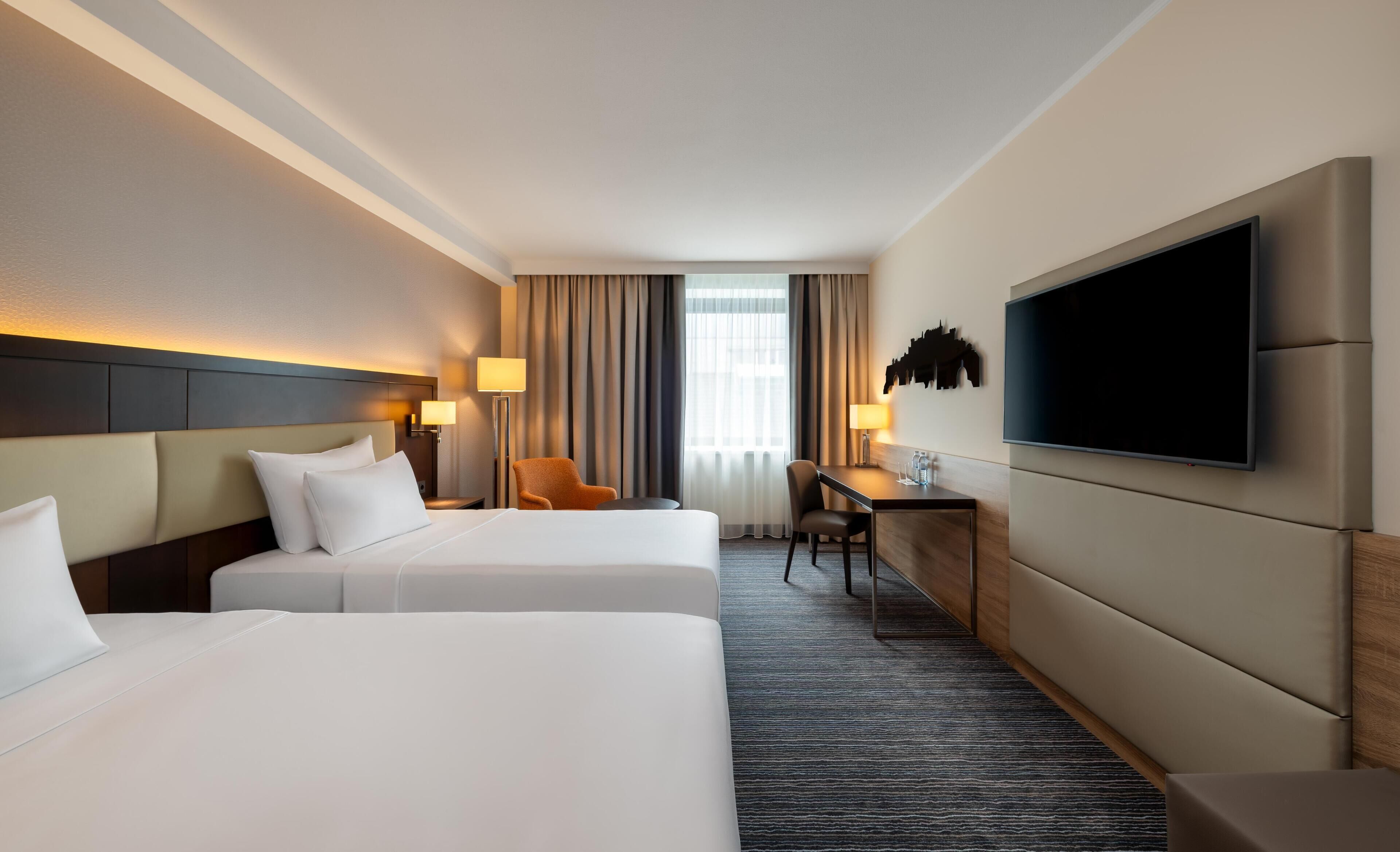 A hotel room with two beds, a desk, a chair, a TV, and a window with curtains.