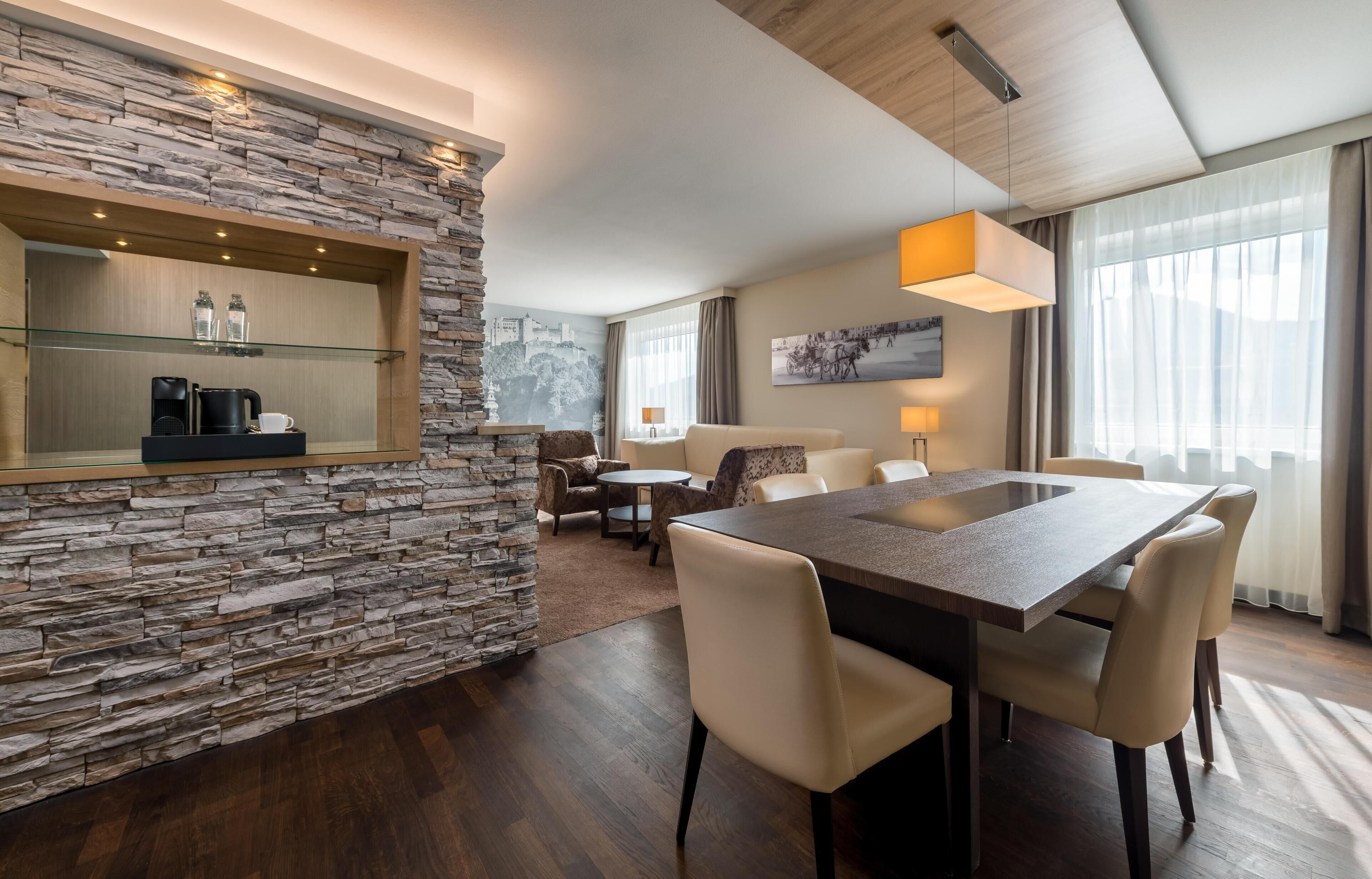 Una habitación de hotel moderna con una pared de acento de piedra, una zona de comedor con una mesa grande y sillas, y un rincón de estar acogedor con un sofá y sillones.