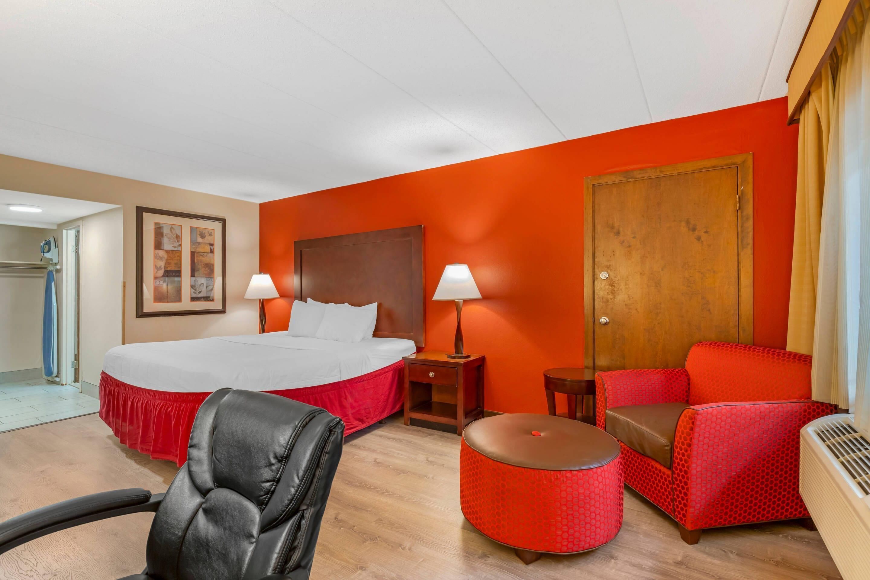 A hotel room with a large bed, a red armchair, a red ottoman, a wooden nightstand, and a wall-mounted heater.