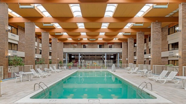 A large indoor swimming pool with lounge chairs and tables, surrounded by brick walls and a high ceiling with skylights. - Clarion Hotel & Conference Center Lansing West (Lansing)