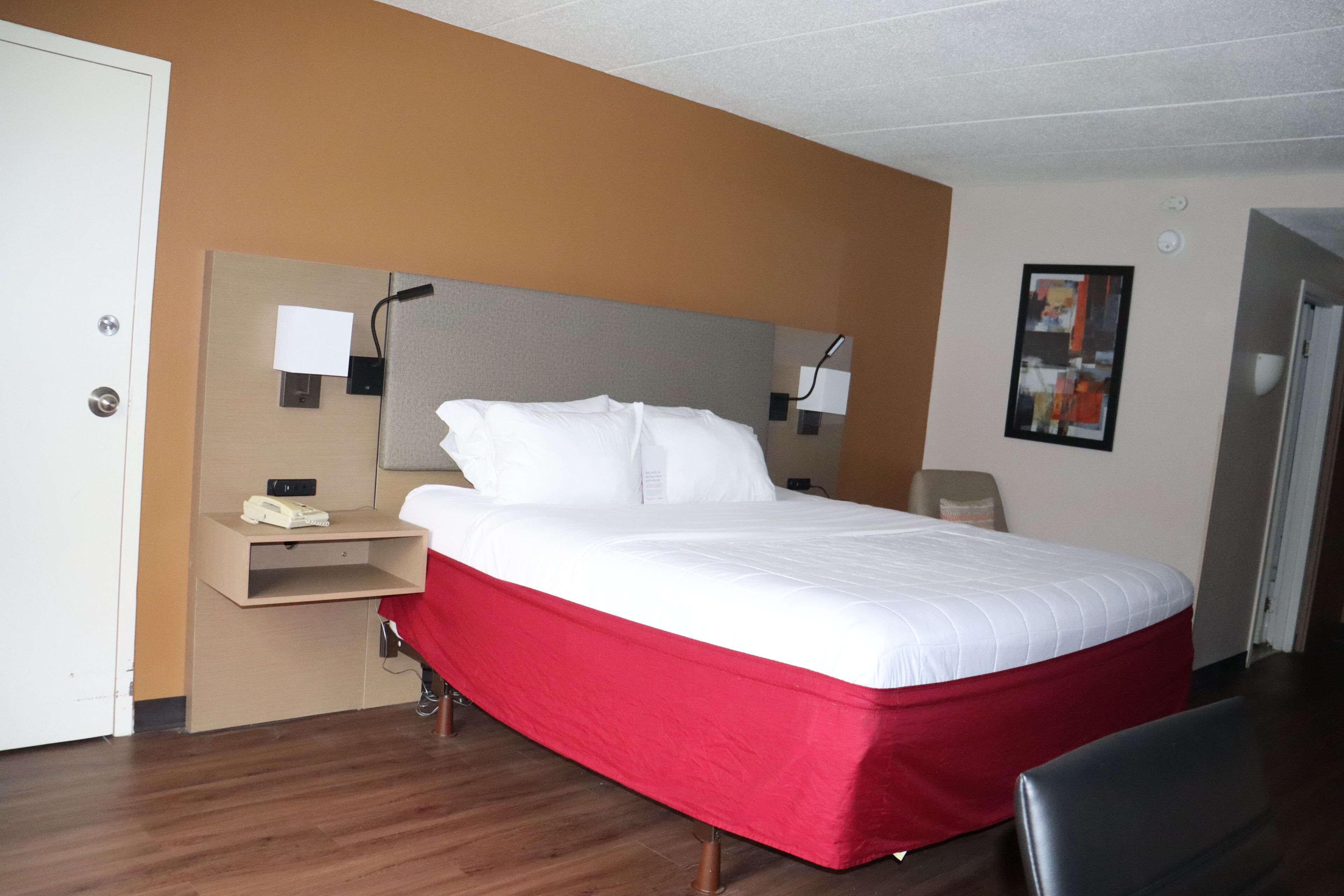 A hotel room with a bed, a desk with a phone, and a wall-mounted lamp.