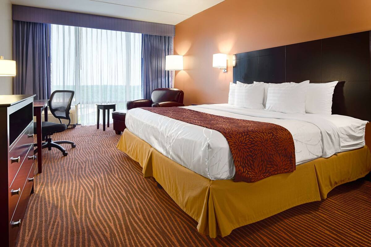 A hotel room with a large bed, a desk with a chair, a small table, and a lamp.