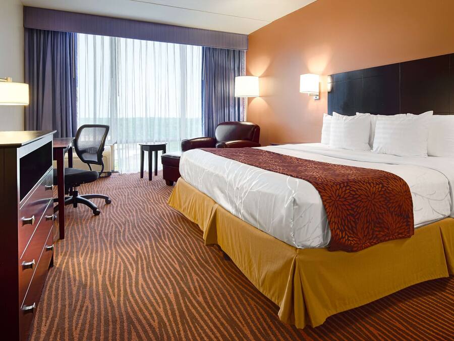 A hotel room with a large bed, a desk with a chair, a small table, and a lamp.