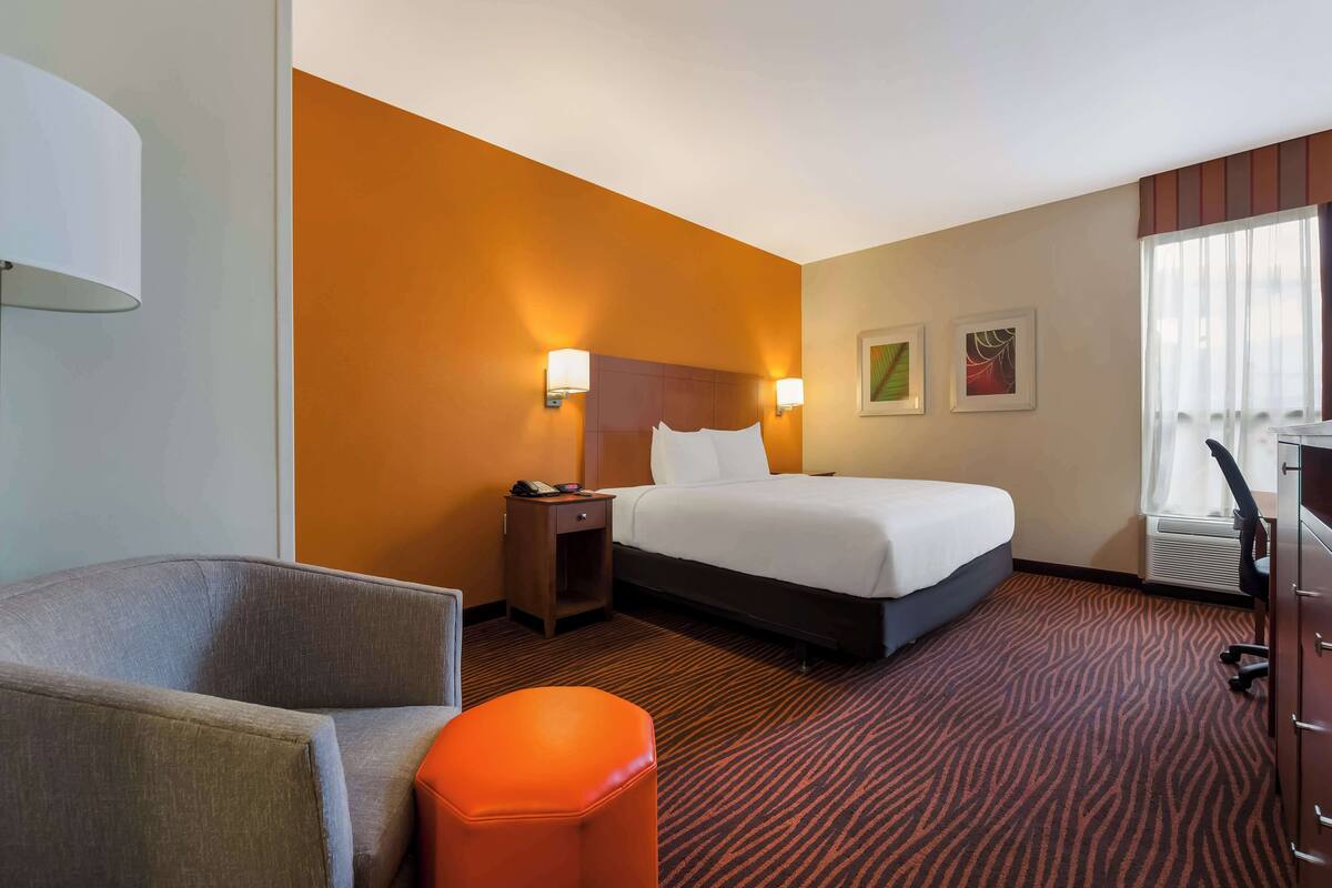 A hotel room with a bed, a chair, an ottoman, a nightstand, and wall-mounted lamps.