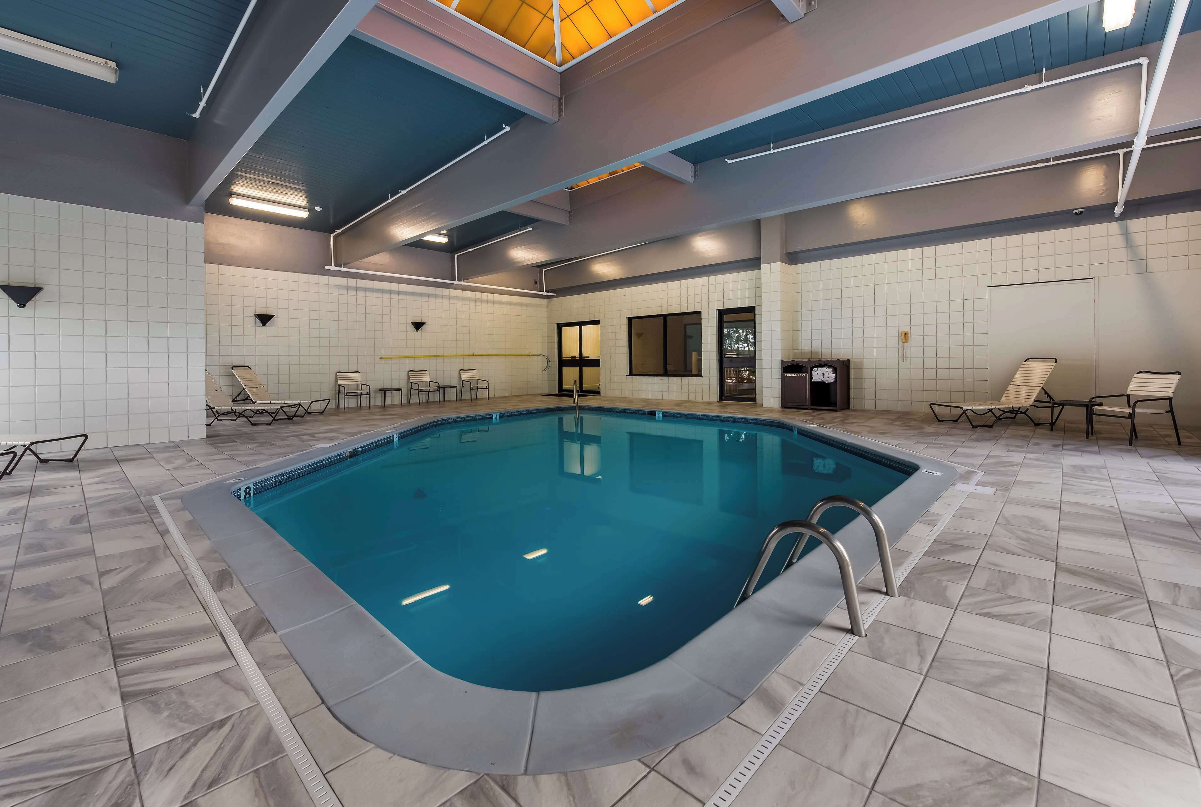 Une piscine intérieure avec un sol carrelé, un escalier en métal et un plafond laissant apparaître les tuyaux et doté d’un système d’éclairage.