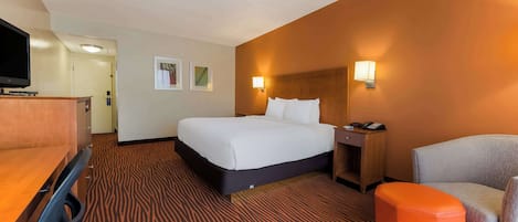 Una habitación de hotel con una cama, un escritorio, una silla, un taburete naranja y un televisor.