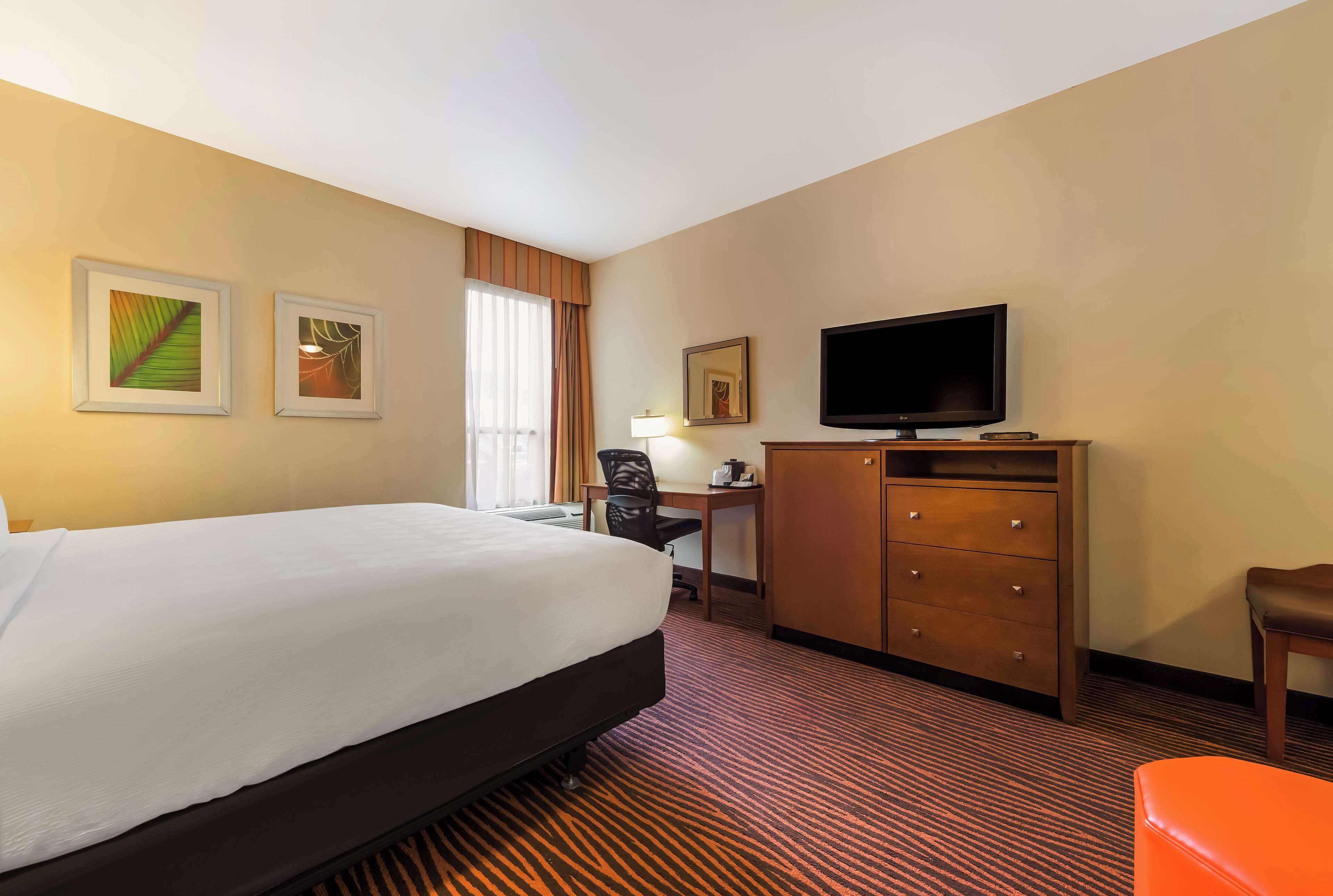 A hotel room with a large bed, a desk with a chair, a television, and a small orange ottoman.