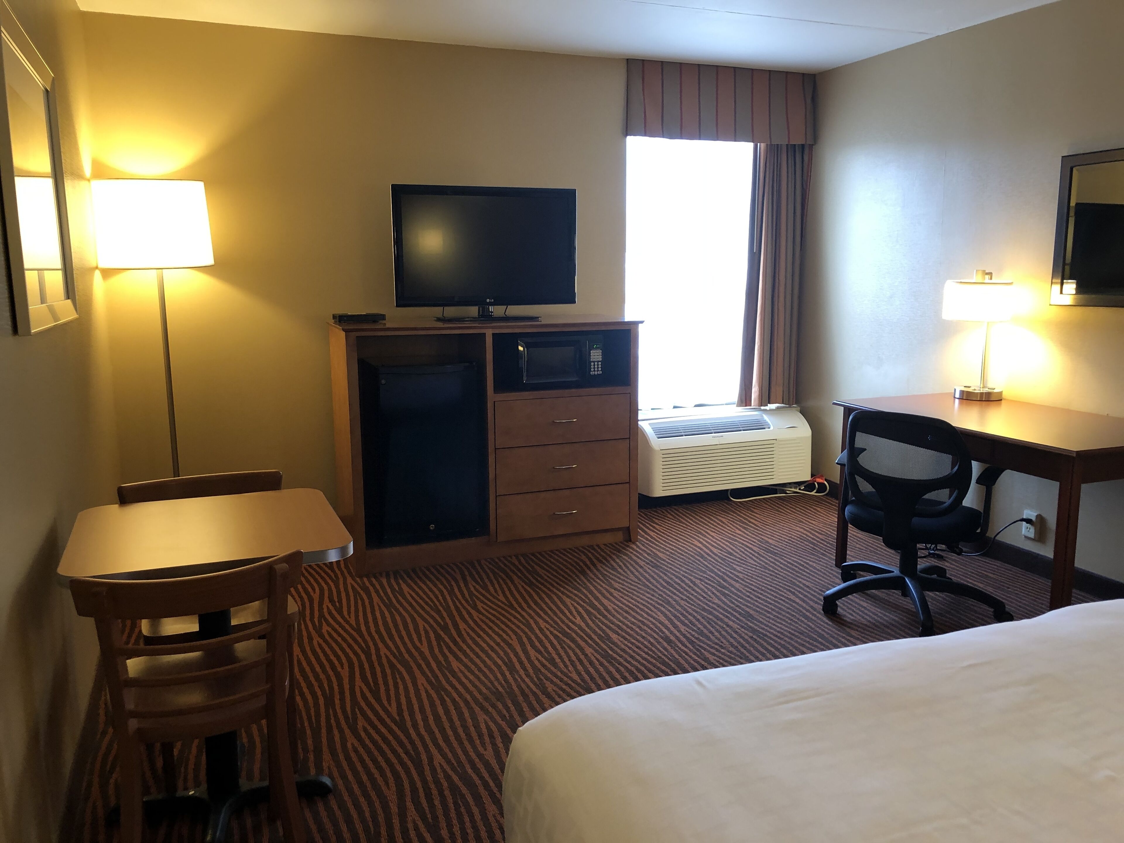 A hotel room with a bed, a desk, a chair, a television, a minibar, and a window with curtains.
