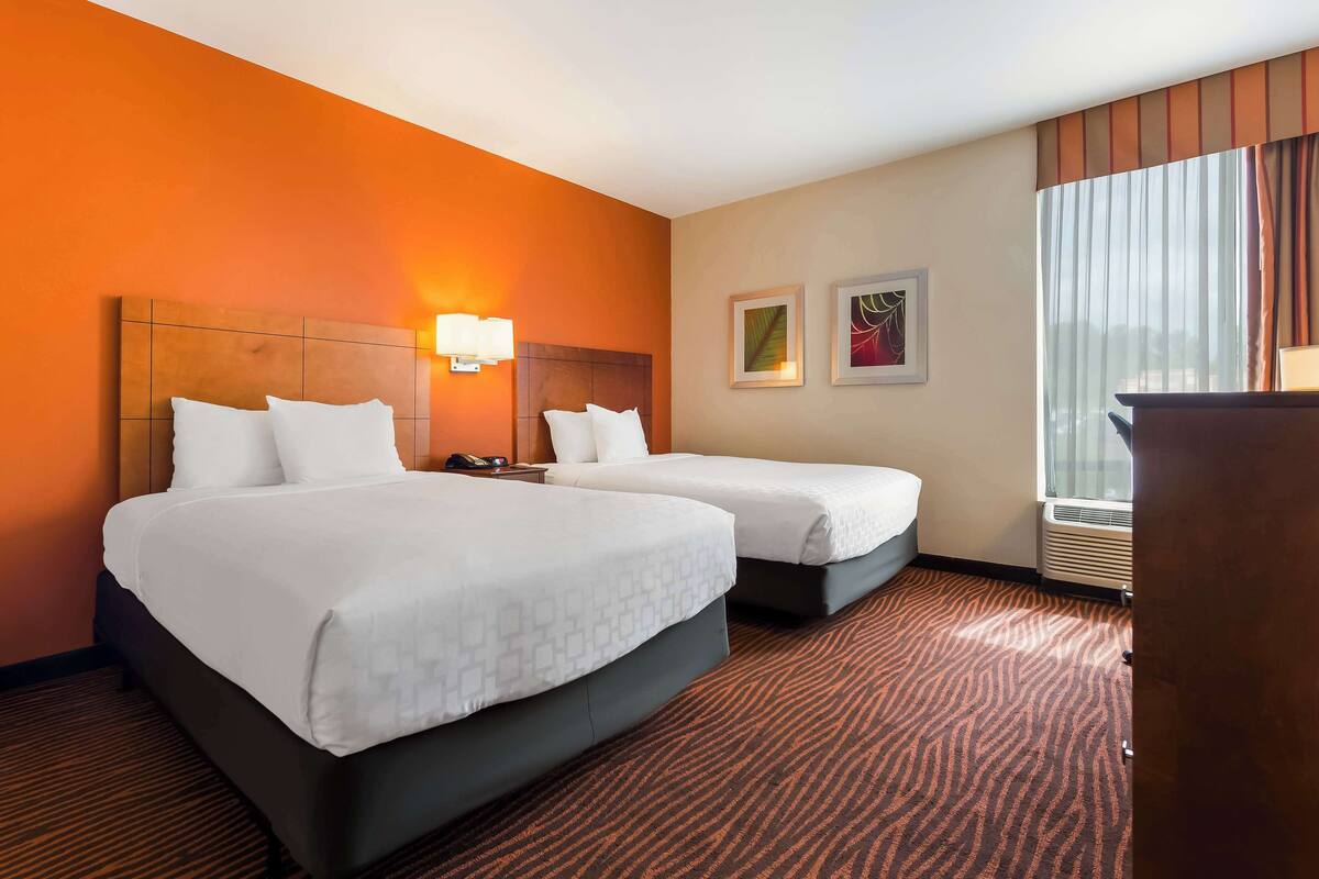 A hotel room with two beds, a wooden headboard, a nightstand, a lamp, framed artwork, and a window with curtains.