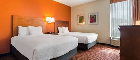 A hotel room with two beds, a wooden headboard, a nightstand, a lamp, framed artwork, and a window with curtains.