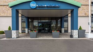 A hotel entrance with a blue and white facade, a sign reading "BW Best Western," and two large planters with red flowers. - Best Western Executive Hotel Of New Haven - West Haven (West Haven)