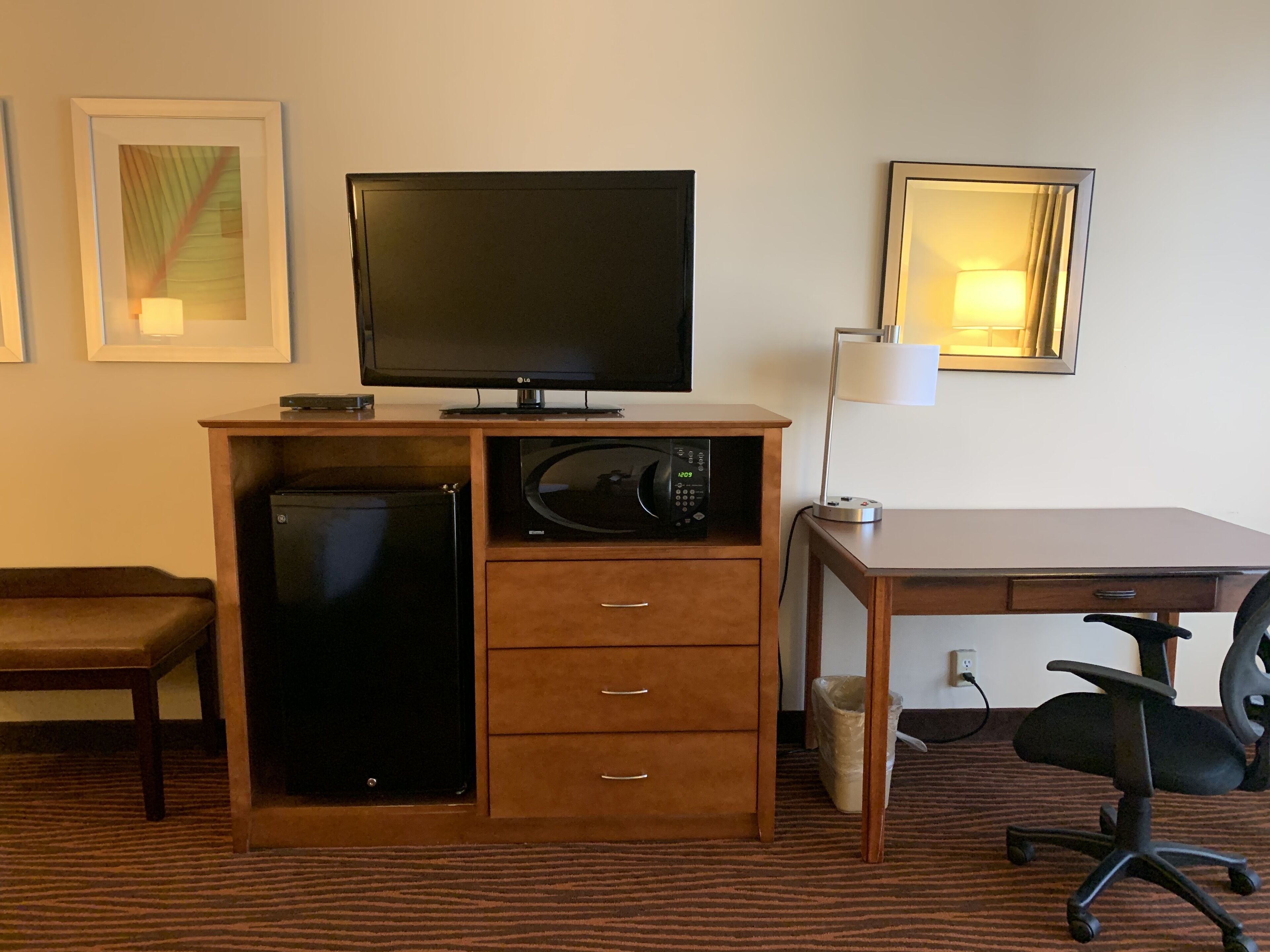 A hotel room with a wooden desk, a television, a microwave, a refrigerator, a chair, and a lamp.