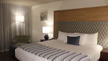 A hotel room with a large bed, a chair, a nightstand with a lamp, and a wall-mounted painting.