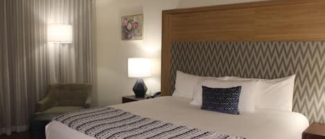A hotel room with a large bed, a chair, a nightstand with a lamp, and a wall-mounted painting.