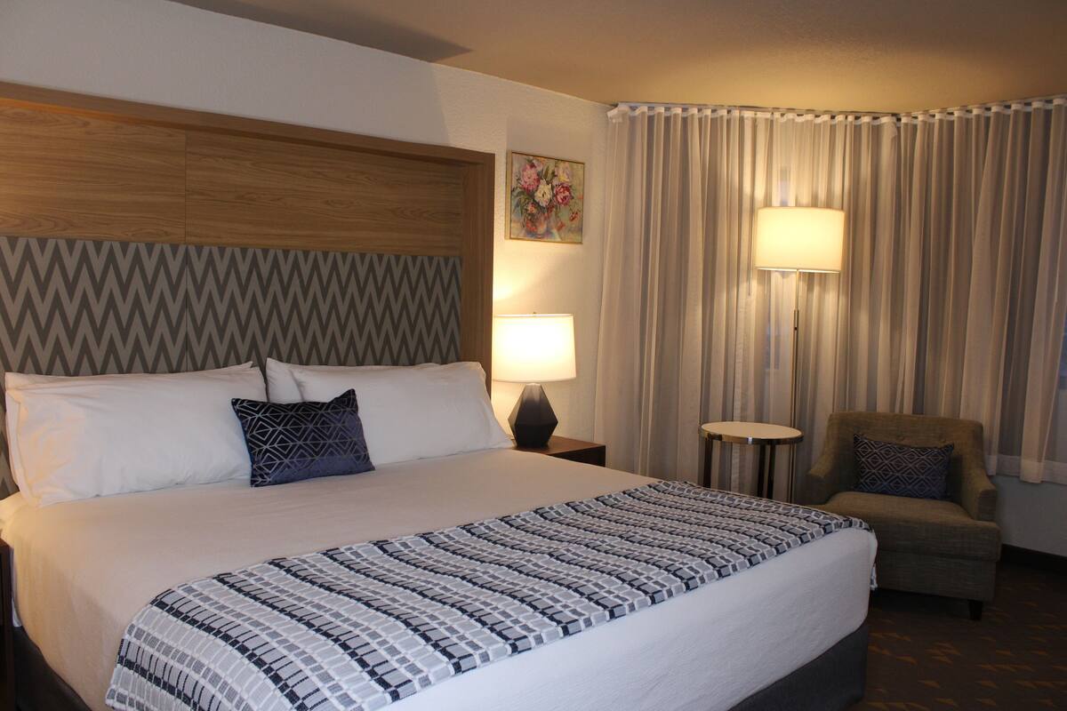 A hotel room with a large bed, a wooden headboard, a patterned bedspread, a nightstand with a lamp, a chair, and a painting on the wall.