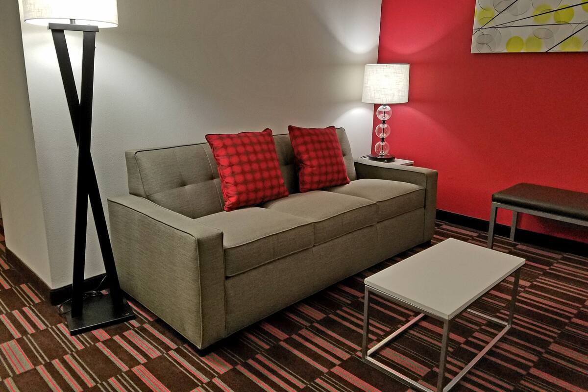 A modern hotel room with a sofa, two red pillows, a side table with a lamp, a small table, and a patterned carpet.