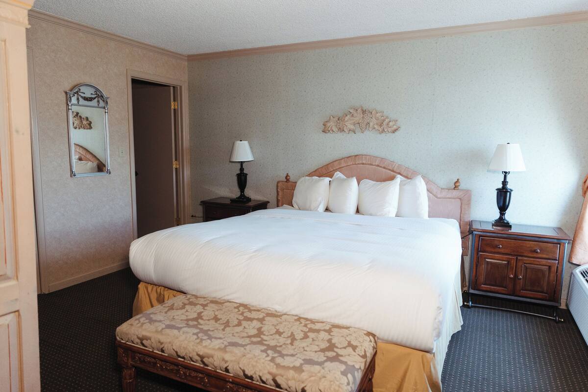 A hotel room with a large bed, a wooden bench, two nightstands with lamps, a mirror, and a decorative wall piece.