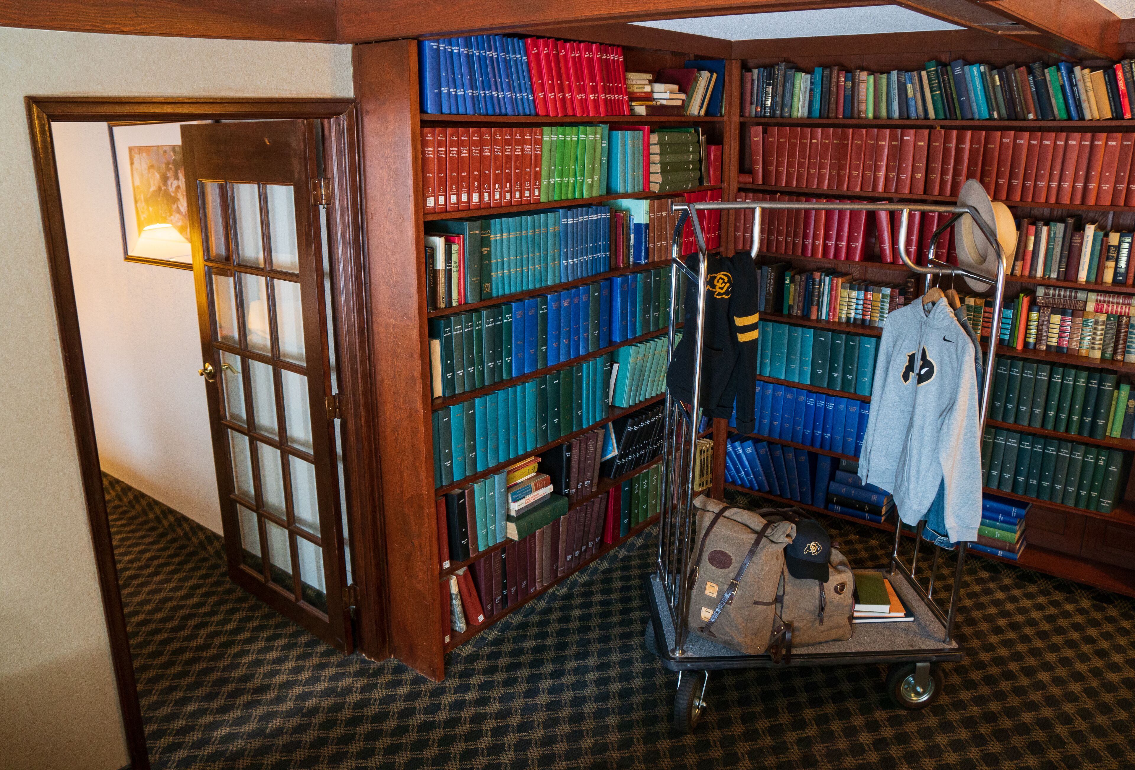 Ett bibliotek med bokhyllor fulla av böcker, en vagn med en jacka och en keps på, och en dörr som leder till ett annat rum.
