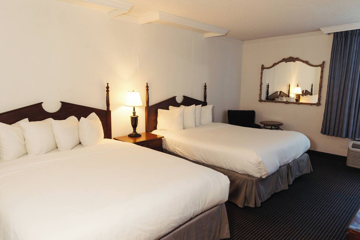 A hotel room with two beds, a nightstand with a lamp, a small table, a mirror, and a window with curtains.