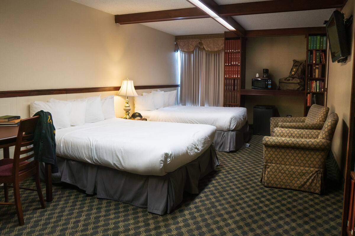 A hotel room with two beds, a desk, a chair, a bookshelf, and a television.